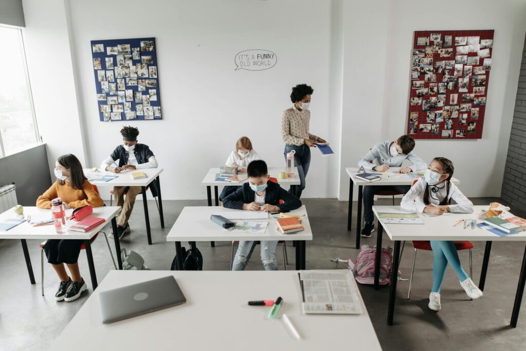 Students studying in a classroom, wearing masks for COVID-19 safety, supervised by a teacher.